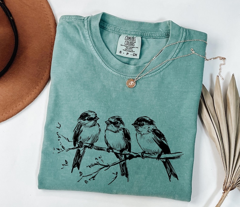 May include: A teal Comfort Colors t-shirt featuring a black illustration of three birds perched on a branch. A silver necklace with a small pendant rests on the shirt. The shirt is folded, with a brown hat visible in the background.