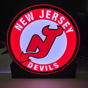May include: A circular, illuminated sign displaying the New Jersey Devils logo. The logo features a red 'N' with horns, set against a white backdrop, surrounded by a red ring with 'NEW JERSEY DEVILS' in white lettering.
