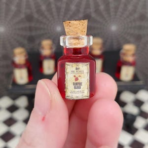 May include: A miniature glass bottle filled with red liquid, labeled "Vampire Blood." The bottle has a cork stopper and a clear plastic collar. Several similar bottles are visible in the background, suggesting a themed collection or display.