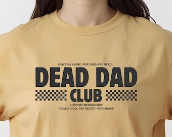 Peut inclure: T-shirt jaune moutarde avec le texte "DEAD DAD CLUB" en lettres noires. Le t-shirt comprend les phrases "LEAVE US ALONE, OUR DADS ARE DEAD!", "LIFETIME MEMBERSHIP" et "REALLY COOL TOP SECRET HANDSHAKE".