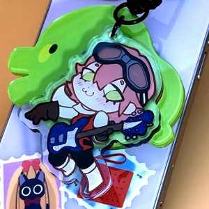 Puede incluir: Un charm de acrílico transparente con un personaje de dibujos animados tocando una guitarra azul, sujeto a un teléfono. El charm tiene una forma verde con forma de dinosaurio detrás. Hay dos imágenes tipo pegatina en el teléfono.