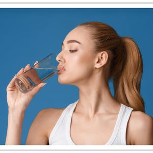 May include: A person drinking water from a clear glass. The person is wearing a white tank top and has their hair pulled back in a ponytail. The background is a solid blue.
