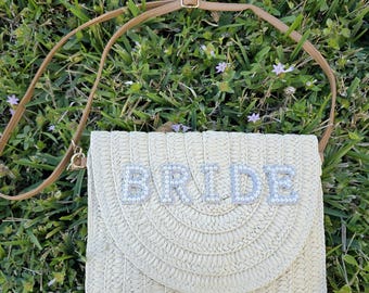 May include: A beige woven clutch with the word "BRIDE" spelled out in silver rhinestones. The clutch has a brown strap and a flap closure. The background is green grass.