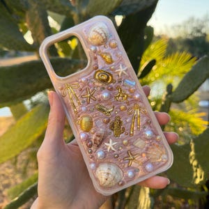 May include: A decorative phone case with a pink background, adorned with seashells, pearls, and gold-colored charms. The charms include a cowboy hat, cactus, and starfish. The case is designed to protect the phone while adding a touch of style.