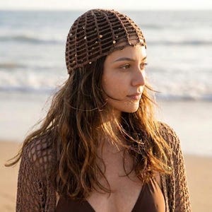 May include: A brown crochet hat with a grid pattern and small sequins. The hat is worn with a matching brown crochet cardigan and a brown bikini top. The background shows a beach and ocean.