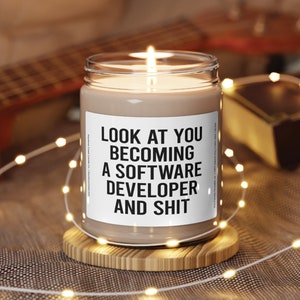 May include: A lit candle in a glass jar with a white label that reads "LOOK AT YOU BECOMING A SOFTWARE DEVELOPER AND SHIT." The candle sits on a wooden coaster, with string lights and a guitar in the background.