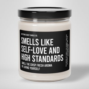 May include: A white soy candle in a glass jar with a black label that reads "Smells Like Self-Love and High Standards" and "Smell the crisp fresh aroma of loving yourself".