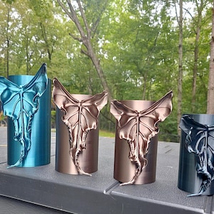 May include: Four cylindrical containers in metallic blue, copper, bronze, and dark gray, each featuring a prominent, three-dimensional moth wing attachment on the front surface, arranged in a row against a natural outdoor backdrop.