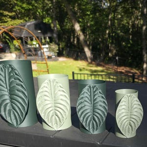 May include: Four cylindrical vases in varying shades of green, each featuring a prominent, raised, textured leaf appliqué on the front. The vases are arranged in a row, alternating between dark forest green and light sage green colors.