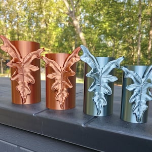 May include: Four cylindrical planters arranged in a row, each featuring a prominent, raised, three-dimensional leaf design on the front. The two planters on the left have a metallic copper finish, while the two on the right are silver.