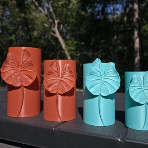 May include: Four cylindrical vases arranged in a row, featuring two terracotta-colored vases on the left and two turquoise-colored vases on the right. Each vase is adorned with a single, raised, textured alocasia leaf design on the front.