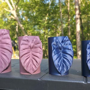 May include: Four cylindrical containers arranged in a row, each featuring a prominent, raised leaf-shaped embellishment on the front. The two containers on the left are mauve, while the two containers on the right are a deep navy blue.