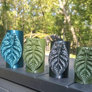May include: Four cylindrical containers, each featuring a prominent, raised, textured leaf emblem on the front. The containers are finished in metallic blue, matte olive green, glossy black, and matte olive green, respectively.