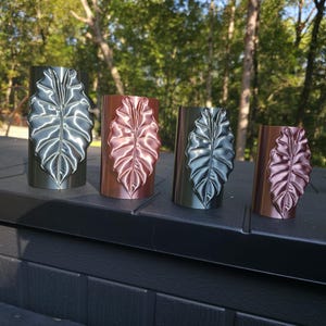 May include: Four cylindrical vases arranged in a row, each featuring a prominent embossed leaf design on the front. The vases alternate between metallic silver and metallic copper finishes, showcasing a smooth, reflective surface texture.