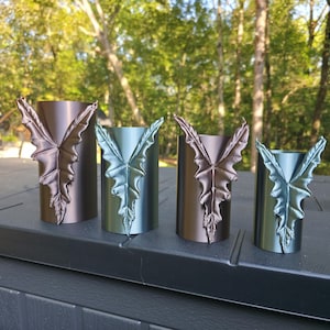 May include: Four cylindrical metal candle holders of varying heights, each featuring a prominent, raised, leaf-shaped embellishment on the front. Two holders have a matte bronze finish, while the other two have a metallic teal finish.