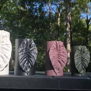 May include: Four cylindrical vases arranged in a row, each featuring a prominent, textured leaf relief on the front. The vases are finished in matte white, black, dusty rose, and dark olive green, with matching leaf designs on each vessel.