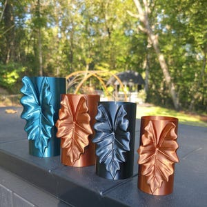 May include: Four cylindrical containers arranged in a row, each featuring a prominent, raised leaf-shaped relief on the front. The containers are finished in metallic blue, copper, black, and copper, showcasing a smooth, reflective surface.