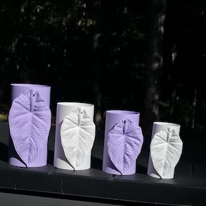 May include: A set of four cylindrical containers in alternating shades of light purple and white. Each container features a single, prominent, textured leaf-shaped appliqué on the front, with the leaf color contrasting against the cylinder base.