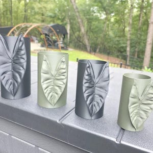 May include: Four cylindrical containers arranged in a row, alternating between matte black and sage green finishes. Each container features a prominent, raised, three-dimensional leaf motif on the front surface, mimicking the shape of an Alocasia leaf.