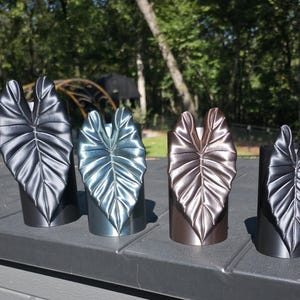 May include: Four cylindrical candle holders, each featuring a prominent, three-dimensional metallic leaf design on the front. The holders are finished in distinct shades of matte black, metallic blue, metallic bronze, and matte black.