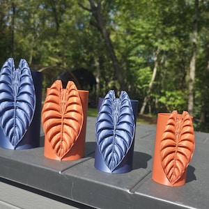 May include: Four cylindrical plant pots arranged in a row, featuring two dark blue and two orange containers. Each pot is adorned with a vertical, three-dimensional relief of a textured, veined leaf design on the front surface.