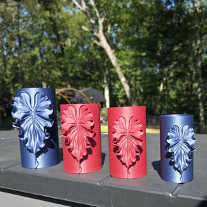 May include: Four cylindrical candle holders arranged in a row. Two are blue and two are red. Each holder features a prominent, three-dimensional embossed leaf design on the front surface, matching the color of the respective cylinder.