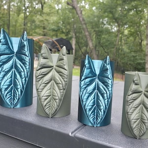 May include: Four cylindrical vases arranged in a row, featuring two metallic blue vases and two matte olive green vases. Each vase is adorned with a single, prominent, three-dimensional leaf relief that matches the color of the cylinder.