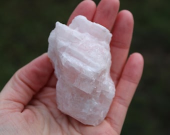 May include: A large, white, and pink selenite crystal with a layered, striated texture. The crystal is held in a hand.