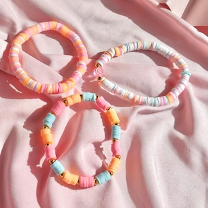 May include: Three colorful beaded bracelets with gold accents. The bracelets are made of pastel pink, blue, and orange beads.