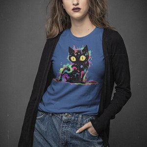 May include: A woman wearing a blue t-shirt with a black cat graphic. The cat is surrounded by colorful paint splatters. She is also wearing a black cardigan and blue jeans.