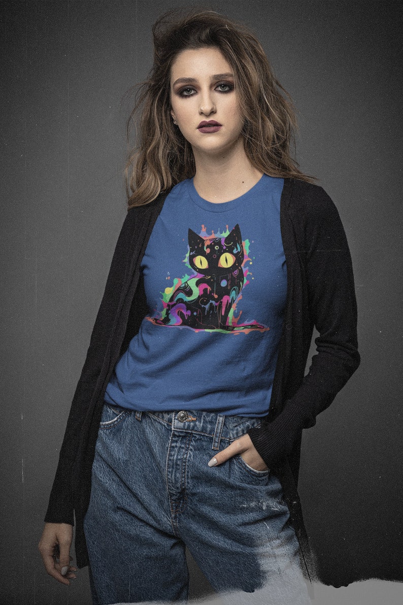 May include: A woman wearing a blue t-shirt with a black cat graphic. The cat is surrounded by colorful paint splatters. She is also wearing a black cardigan and blue jeans.