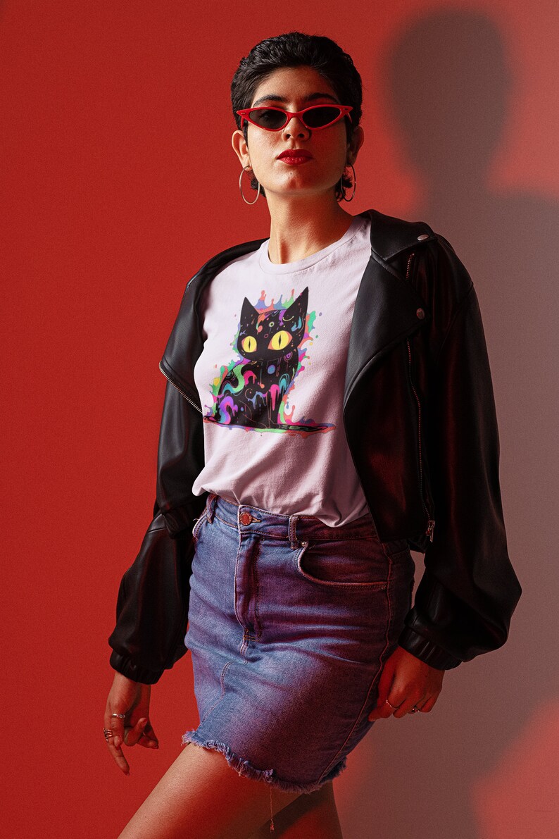 May include: A woman wearing a white t-shirt with a black cat graphic featuring colorful paint splatters. The cat has yellow eyes and a psychedelic design. She is also wearing a black leather jacket, a denim skirt, and red sunglasses.