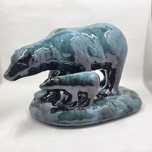 Puede incluir: Una escultura de cerámica de un oso grande de pie sobre un osezno. La pieza presenta un esmalte brillante y variado en tonos azul verdoso profundo, azul oscuro y negro, creando un efecto jaspeado sobre la superficie lisa y reflectante.