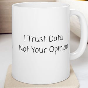 Può includere: Tazza in ceramica bianca con la scritta nera "I Trust Data, Not Your Opinion". La tazza è su un sottobicchiere beige. Lo sfondo comprende un tagliere in legno, una ciotola bianca con frutta e un contenitore in legno.