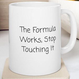 May include: White ceramic mug with the text "The Formula Works, Stop Touching It" in black. The mug sits on a beige coaster. The mug has a curved handle and is in a kitchen setting.