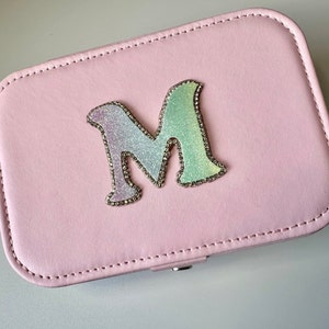 May include: A pale pink rectangular jewellery box with a stitched border. The box features a large, glittery letter "M" in shades of purple, green, and silver, outlined with small, sparkling rhinestones. The box is closed.