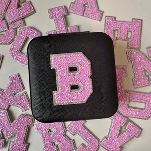 May include: A black jewellery box with a pink sparkly letter B on the front. The box is surrounded by pink sparkly letters.