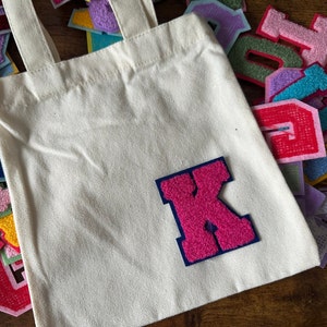 Könnte beinhalten: Eine weiße Canvas-Tragetasche mit einem pinkfarbenen Filz-Buchstaben K-Patch. Der Buchstabe ist dunkelblau umrandet. Die Tasche ist von einer Vielzahl bunter Filzbuchstaben umgeben.