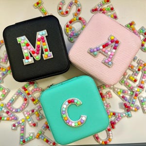 May include: Three square jewellery boxes with different coloured lids. The lids are black, pink and turquoise. Each lid has a large letter on it made of colourful beads. The letters are M, A and C.