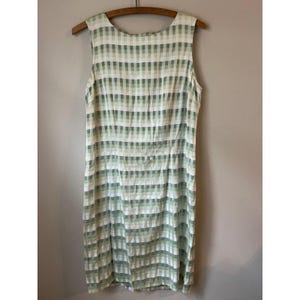 May include: Sleeveless dress with a green and white checkered pattern. The dress is hanging on a wooden hanger. The fabric has a soft, lightweight appearance, suitable for warm weather. The dress is knee-length.