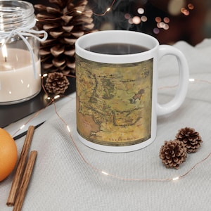 May include: A white ceramic mug featuring a detailed, vintage-style map of Middle-earth printed on its exterior. The map displays various geographical regions, mountain ranges, and text labels in a fantasy script, set against a muted, aged parchment background.