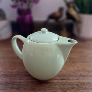 May include: A pale mint green ceramic teapot with a rounded body, a curved handle on the left side, and a short, wide spout on the right. The teapot features a matching removable lid with a small knob handle centered on top of the smooth surface.