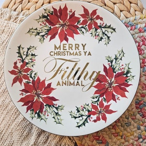 Könnte beinhalten: Ein weißer Keramikteller mit schwarzem Rand, verziert mit roten Weihnachtssternen, grünem Stechpalmenlaub und goldfarbenem Schriftzug. Der Teller trägt den Text "MERRY CHRISTMAS YA Filthy ANIMAL".