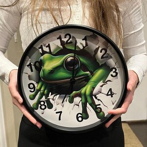 May include: A black and white clock with a green frog breaking through the face. The clock has Roman numerals and a black background.