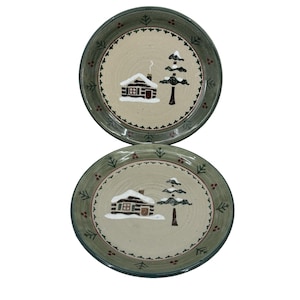 May include: Two round ceramic plates featuring a beige center with a hand-painted brown log cabin and a dark green pine tree covered in white snow. The wide, sage green rim is decorated with a repeating pattern of small pine trees and red berries.