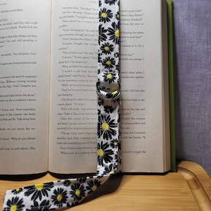May include: A black and white daisy patterned fabric bookmark with a silver ring. The bookmark is placed on an open book with text about a cooking show.