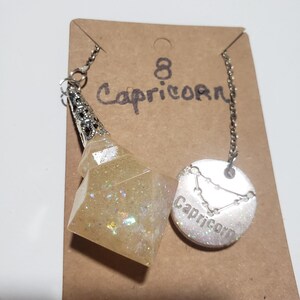 May include: A silver chain necklace with a white glitter resin pendant shaped like a pyramid and a silver charm with the zodiac sign Capricorn and the word "Capricorn" inscribed on it. The necklace is displayed on a brown card with the number "8" and the word "Capricorn" written on it.