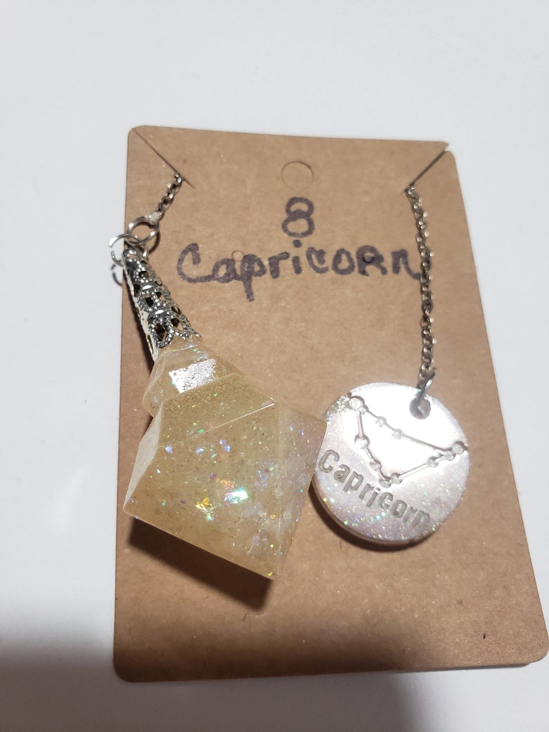 May include: A silver chain necklace with a white glitter resin pendant shaped like a pyramid and a silver charm with the zodiac sign Capricorn and the word "Capricorn" inscribed on it. The necklace is displayed on a brown card with the number "8" and the word "Capricorn" written on it.