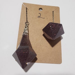 May include: A pair of dangle earrings with a dark purple glitter finish. The earrings are shaped like a teardrop and a cube. The earrings are hanging from a brown card with the number "2" written on it.