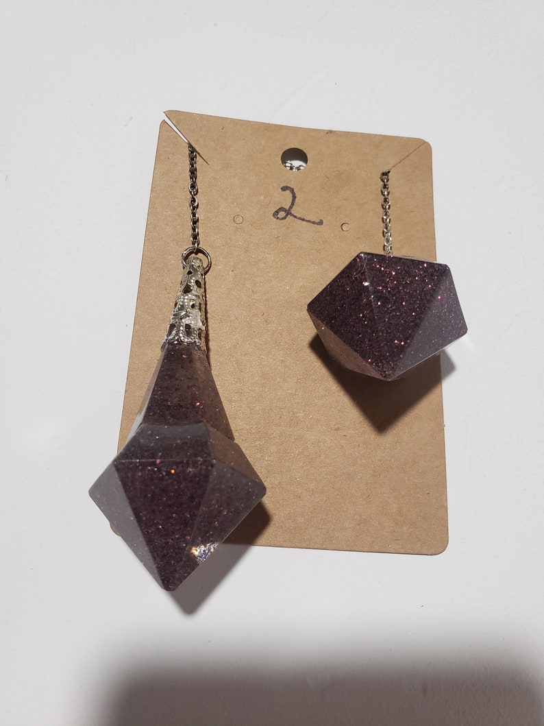 May include: A pair of dangle earrings with a dark purple glitter finish. The earrings are shaped like a teardrop and a cube. The earrings are hanging from a brown card with the number "2" written on it.